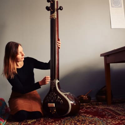 Laura Naukkarinen sitter på knä och spelar på ett indiskt stränginstrument i ett rum med turkisk matta och blå vägg.