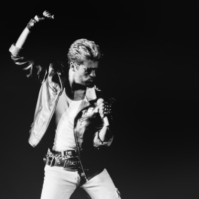 Mustavalkoisessa kuvassa laulaja George Michael lavalla nahkatakissa ja valkoisissa farkuissa, vasemmassa kädessä mikrofoni, oikea käsi nostettuna nyrkissä pään yläpuolelle.