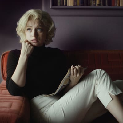 Marilyn Monroe (Ana de Armas) sitter i en soffa med en tidning i handen och ser förbryllad ut.