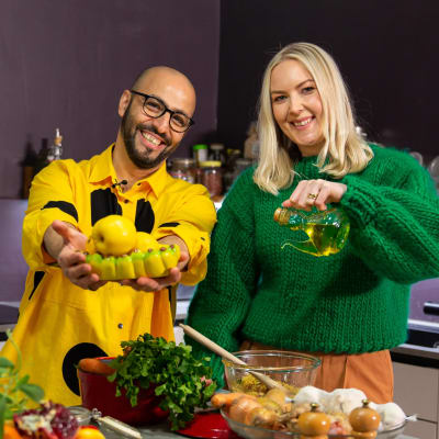 Mies keltaisessa paidassa ja nainen vihreässä paidassa katsoo kameraan. Edessä pöydällä eri ruoka-aineita, kuin sipulia, yrttejä ja oliiviöljyä.