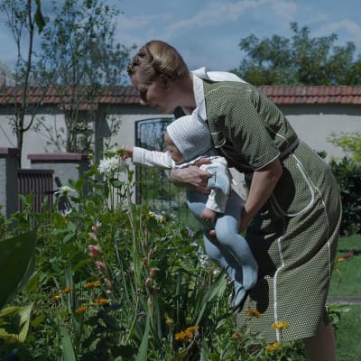 En kvinna med ett litet barn i famnen lutar sig fram över en vacker blomma i en rabatt.