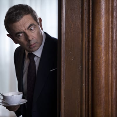 Johnny English tjuvlyssnar bakom en dörr.