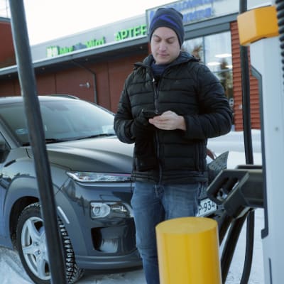 En man står med sin mobiltelefon vid en snabbladdningsstation. I bakgrunden syns hans elbil.