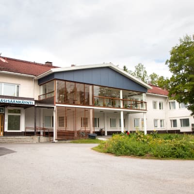 Ruoveden terveyskeskus kuvattuna pääovien edestä. Keskellä pihaa kukkaistutuksia, rakennuksessa muun muassa iso lasitettu parveke.