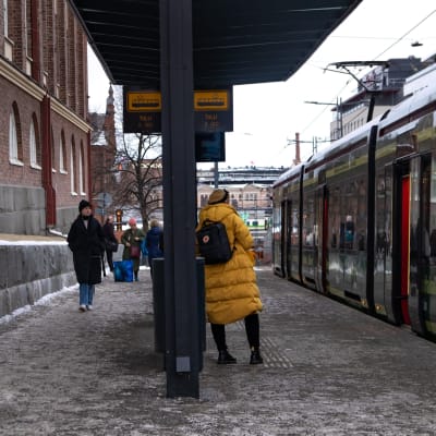 Ihmisiä tamperelaisella ratikkapysäkillä. Keltatakkinen nainen nojailee pysäkkiin, ja vieressä ratikka tekee lähtöään.