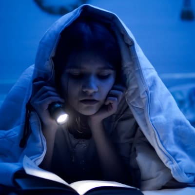 En liten flicka läser med ficklampa under täcket i ett mörkt rum.