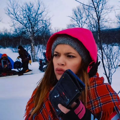 Hilda Länsman pitää puhelinta kädessään Inarin disko -kuvauksista lumisessa maisemassa. Taustalla näkyy sininen teltta ja kaksi ihmistä.