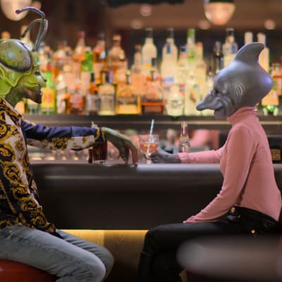 Två personer utklädda i masker är på en första dejt på en bar. Till vänster en man utklädd till en grön insekt och till höger en kvinna utklädd till en fisk..