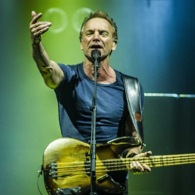 Sting sträcker ut ena armen, har en elbas kring halsen och sjunger i en mikrofon som är i en mikrofonställning.
