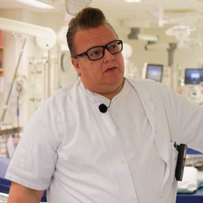 En vitklädd man med svarta glasögon och högt hår står i ett sjukhusrum.