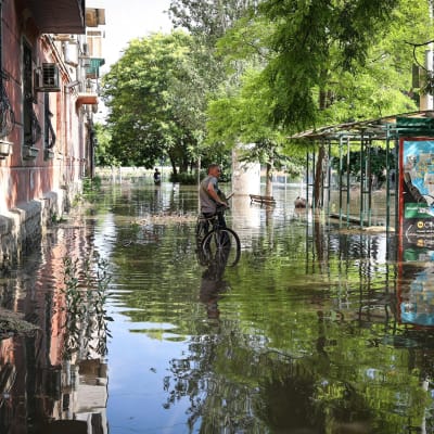 En person lutar mot sin cykel på gatan. Det finns översvämmningsvatten upp till vristrena på gatan. 