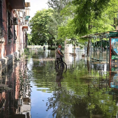 En person lutar mot sin cykel på gatan. Det finns översvämmningsvatten upp till vristrena på gatan. 