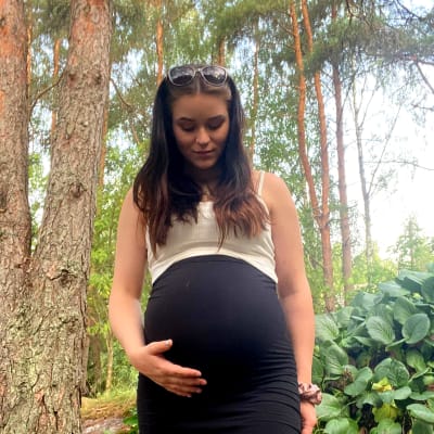 En ung gravid kvinna står i skogskanten och tittar på sin mage.