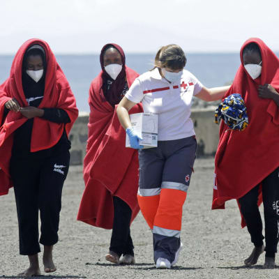 Tre kvinnor i röda filtar eskorteras av en hjälparbetare på en strand