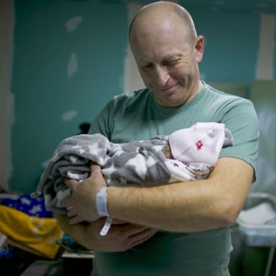 En man håller i en liten bebis i famnen. Han tittar ner på bebisen och ler.