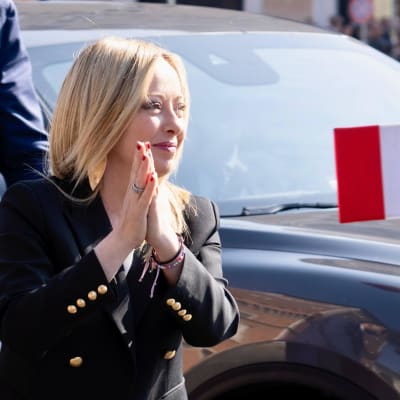 Mustajakkuinen vaalea nainen painaa kämmenensä yhteen mustan auton edessä ja hymyilee oikealle päin. Taustalla auton konepellillä on Italian lippu.