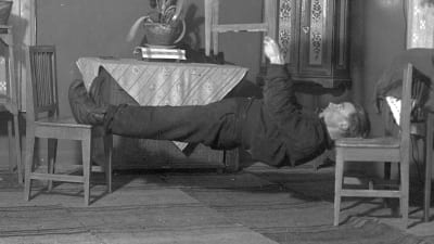 En man leker "ligga lik", där man ska hålla kroppen så styv som möjligt medan man ligger mellan två stolar. Bilden är tagen 1937 i Närpes, Kalaks.