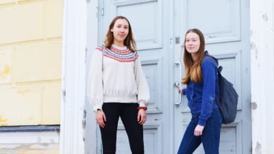 Två unga kvinnor står på trappan till Lovisa Gymnasium, ett gult trähus med blå ytterdörr. Den ena av dem håller i dörrhandtaget.