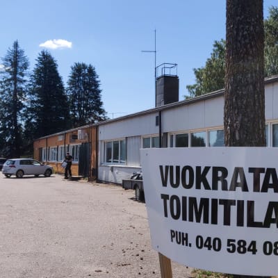 I högra hörnet på bilden syns en skylt där det står på finska att affärsutrymmen uthyres. Bkom skylten synns ett lågt långt hus.