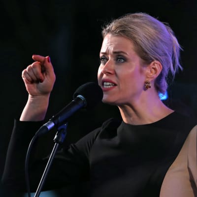 Kvinna med blont hår håller upp fingret medan hon talar.