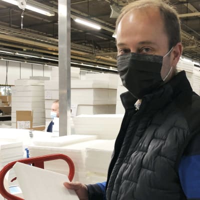 Bild på Peter Nordlund, iklädd svart munskydd tillverkat i hans fabrik. Han håller i ett kassettfilter och står inne i den fabrikshall som tillverkar dem.