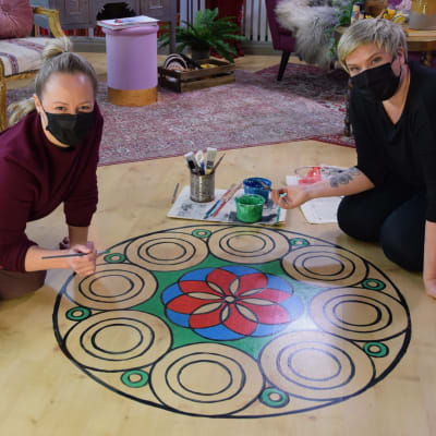 Två blonda kvinnor med ansiktsmasker som sitter på golvet i ett rum och målar en stor mandala på laminatgolvet.