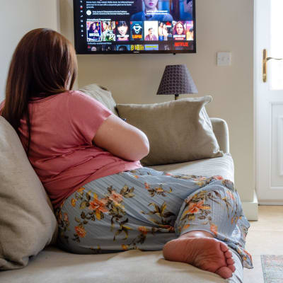 En kvinna sitter i en soffa och tittar mot en tv där man kan se Netflix utbud.