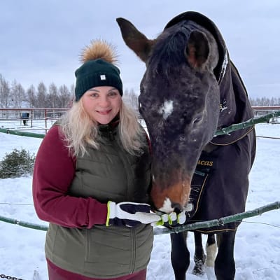 Nuori vaaleahiuksinen nainen seisoo hevosaitauksen edessä, maassa on lunta. Aitauksen kalterien yli kurottaa iso ruskea hevonen, joka haistelee naisen kämmentä.