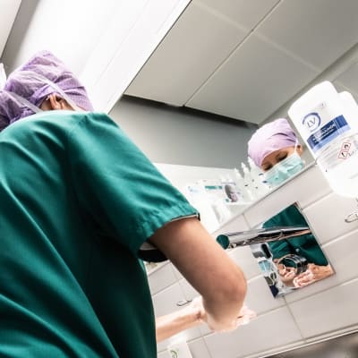 En kvinna med håret bakbundet och skyddat tvättar händerna innan hon går in i en operationssal. Hon har på sig gröna kirurgkläder.