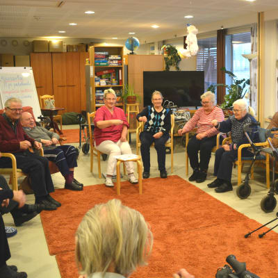 Många äldre personer sitter inomhus på stolar i en ring. De ser glada ut och har händerna i luften.