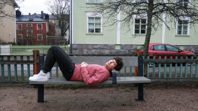 Kasper Strömman ligger på en parkbänk med fina trähus i bakgrunden i Trävallgård i Helsingfors.