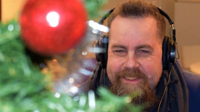 Stan Saanila tittar fram bakom julgran i förgrunden. Han har hörlurar på huvudet och ler.
