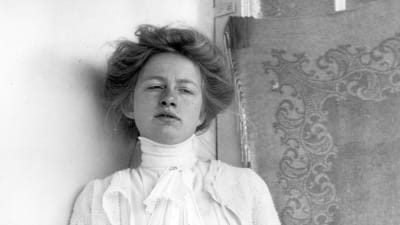 Edith Södergran mot tapet i lång vit klänning. Måttband i handen. Uppsatt hår. blicken snett nedåt.