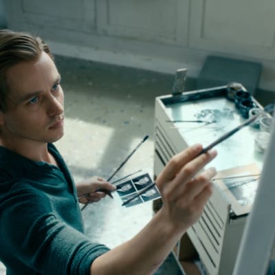 Kurt Barnert (Tom Schilling) maalaa ateljeessaan. Kädessä hänellä on mallina neljän passikuvan arkki.