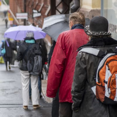 Människor som står i en brödkö. Flera har paraply efetrsom det regnar. Personerna står med ryggen mot kameran.