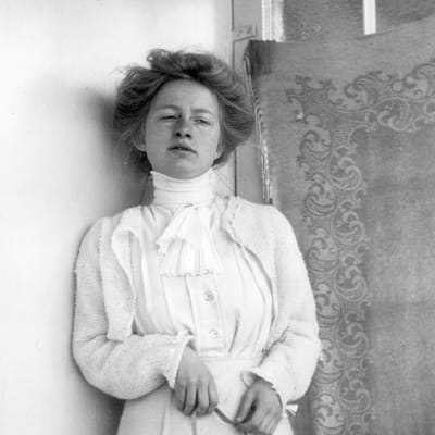 Edith Södergran mot tapet i lång vit klänning. Måttband i handen. Uppsatt hår. blicken snett nedåt.