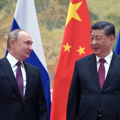 Två äldre män i mörk kostym och violett slips tittar snett på varandra. I bakgrunden Kinas och Rysslands flaggor.