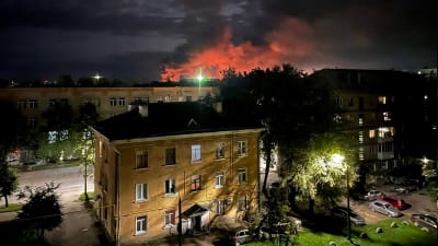 Ett gult trevåningshus i sten och i bakgrunden röd brandrök som stiger mot natthimlen. 