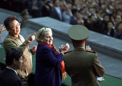 två män i uniformer och en kvinna i lila jacka och orange halsduk klappar händerna.