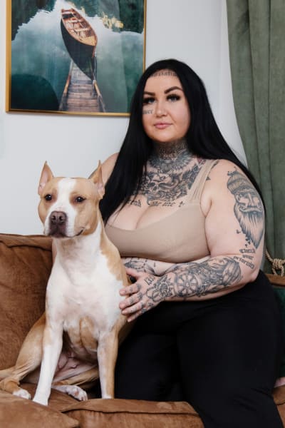 Tummahiuksinen, isokokoinen, tatuoitu nainen istuu sohvalla pitbullin kanssa ja katsoo kameraan. Myös koira näyttäisi katsovan kameraan ja ikään kuin hymyilevän.