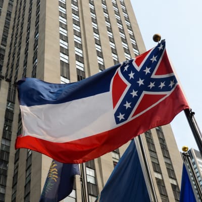 En bild på Mississippis flagga. Uppifrån och ner har den tre ränder i blått vitt och rött. Till höger på flaggan är det ett fyrkantigt rött fält med ett blått kryss över med vita stjärnor - den såkallade sydstatssymbolen.
