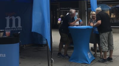 Tre personer diskuterar vid ett bord med Moderaternas logo.