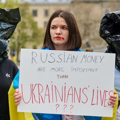 Kvinnor med blodiga kläder och svarta sopsäckar på huvudet demonstrerar i Berlin mot Tysklands ovilja att bojkotta rysk olja och gas. En av kvinnorna är iklädd en ukrainsk flagga och håller upp en skylt med texten "Ryska pengar är viktigare än ukrainska liv?". 