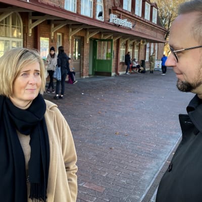 Maakuntajohtaja Anna-Mari Ahonen ja kaupunginjohtaja Olli-Poika Parviainen keskustelevat Hämeenlinnan juna-aseman ratapihalla, punatiilinen asemarakennus taustanaan.