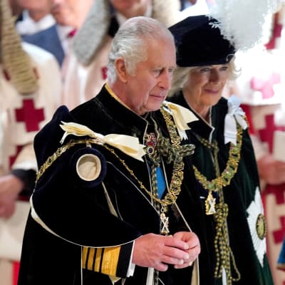 Laajassa puolikuvassa kuningas Charles III ja kuningatar Camilla kävelevät juhla-asuissa ihmisten ohi.