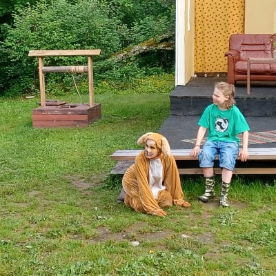 En liten pojke och en person, utklädd till en hund, sitter på en bänk framför ett hus.