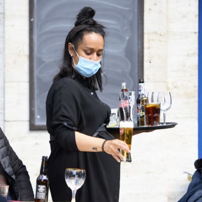 En kvinnlig servitör håller i en bricka med öl på en uteservering. Hon har munskydd på sig.