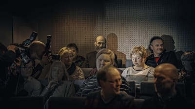 Folk sitter i ett auditorium. Ett par fotografer tar bilder av en kvinna.