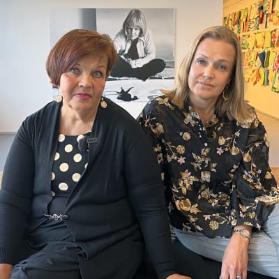 Helena Lindstén ja Hanna Mamia-Walther istuvat Riihimäen taidemuseon penkillä, taustalla heidän välissään valokuvasuurennos Maija Isolasta.