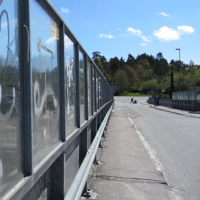 En bro med körfil och trottoar samt skyddsräcken av genomskinlig plast som är klottrat. Sommartid, solen skiner. På bron syns några fotgängare på avstånd.  Under bron skymtar järnvägsspår och ett tåg samt parkerade bilar.